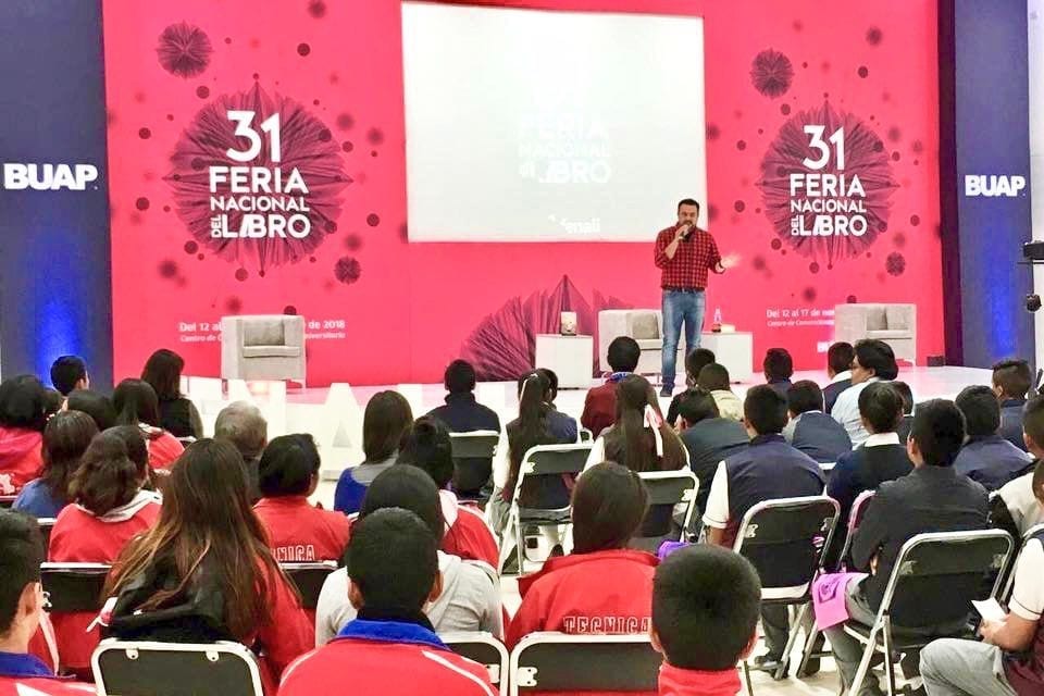 Arturo Ordorika en el escenario de la 31 Feria Nacional del Libro de la BUAP, micrófono en mano, frente a un auditorio lleno de estudiantes