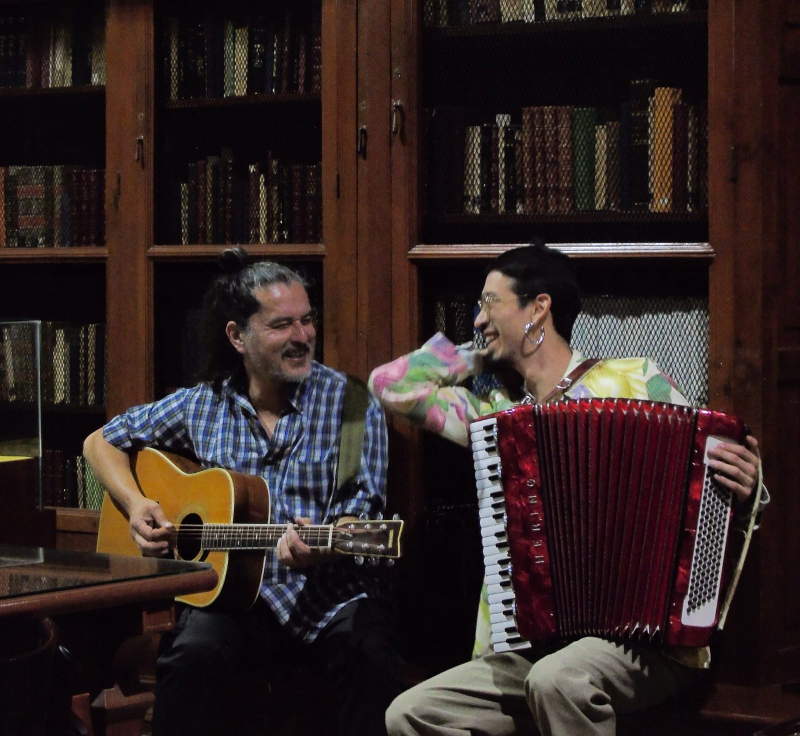 Félix Guerrero, con guitarra, y Carlos Marrufo, con acordeón rojo, riéndose frente a una biblioteca de madera