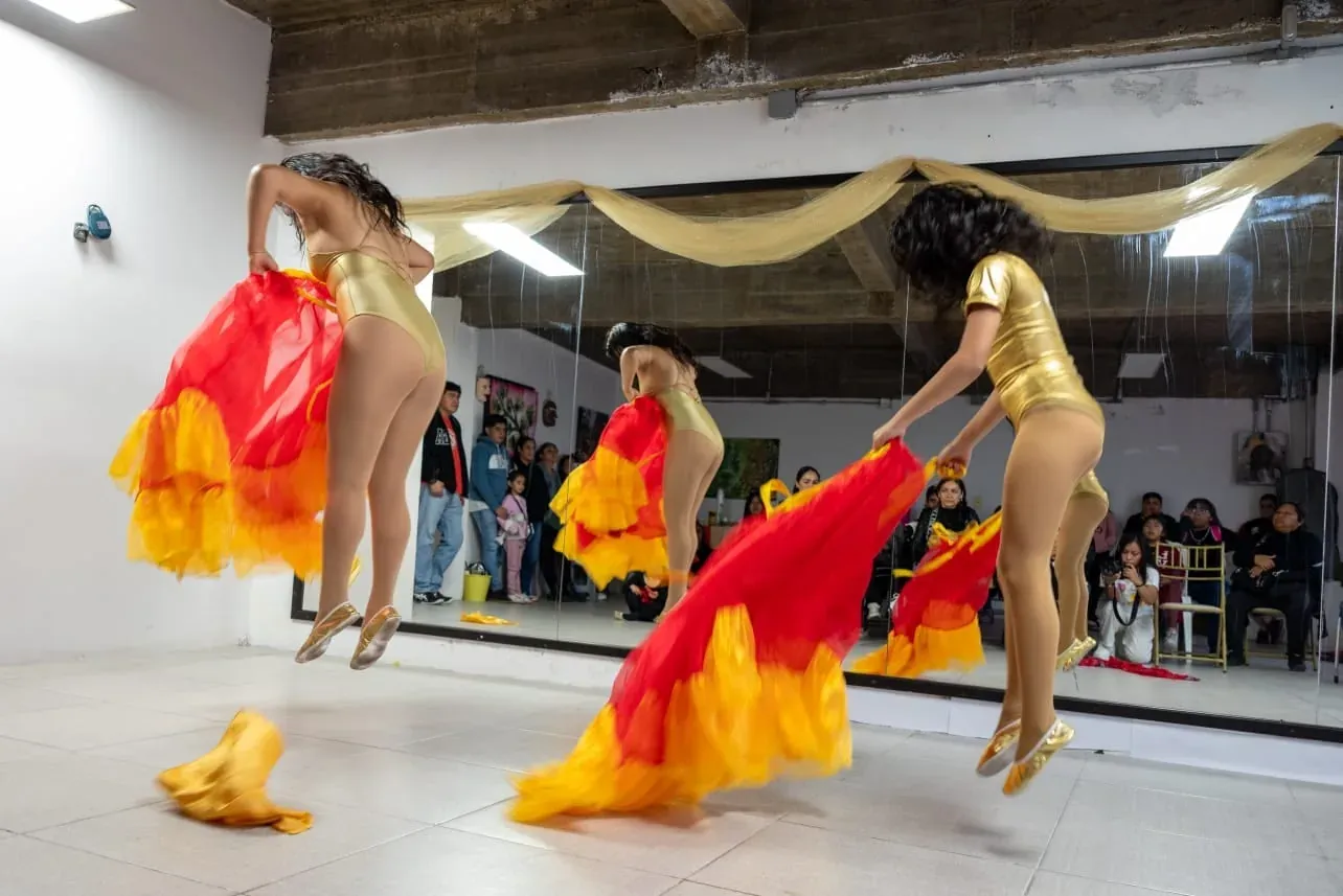 Ángeles Rodríguez and Alis in mid-air, in red and orange skirts and gold costumes, in front of the mirror of the Ollin studio with the audience on its feet