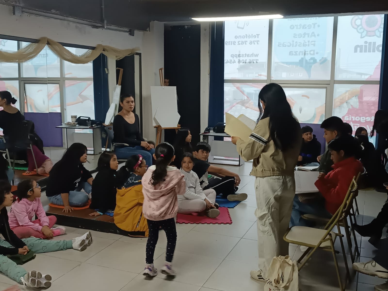 Una clase de Ollin: niñas y niños sentados en el piso del salón de espejos escuchan a una alumna que lee de pie