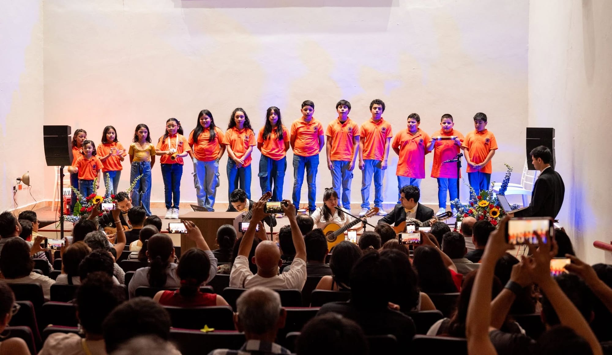 El coro de Ollin Cuauchinanco, niñas y niños de playera naranja, canta en un escenario ante un público que levanta los celulares